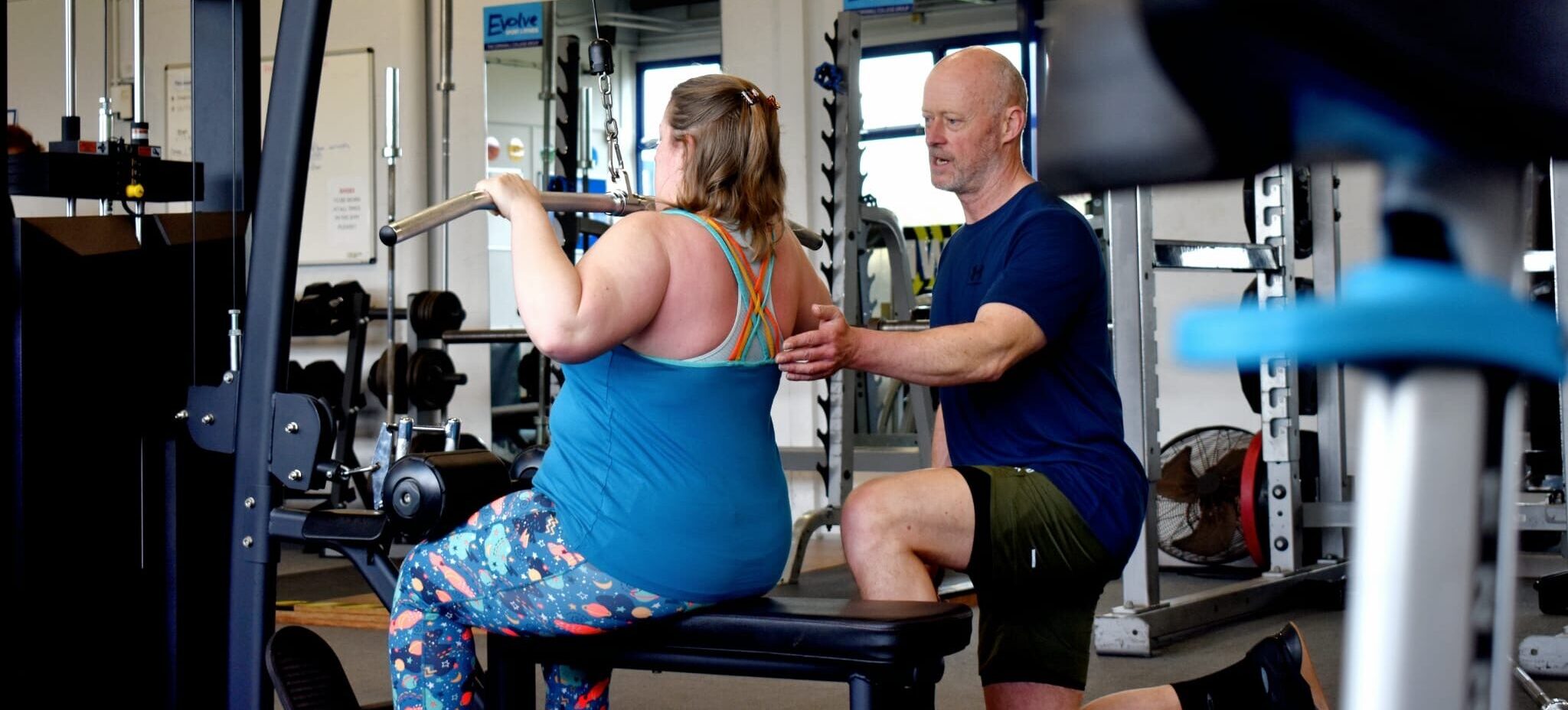 Photograph of M:Power owner training a client in a gym, showing correct posture and technique on a lateral pulldown resistance machine.